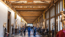 The Uffizi Gallery museum in Florence, Italy, with people walking through the hallways looking at sculptures and paintings on display. the ceiling is painted with frescoes. there's an art gallery-like atmosphere with bright natural light coming from large windows on both sides of the hallway