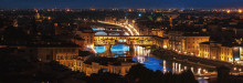  panoramic view of the city at night, with the famous bridge in florence visible from above. the river is flowing through it, and there's an illuminated bridge that connects to buildings on both sides. on one side of the photo, you can see tall buildings, while the other half shows greenery and trees. there are cars driving along the road near the water, adding movement to the scene.